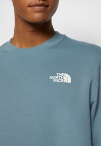 Gros plan sur une personne portant un sweat-shirt col rond bleu clair avec un logo blanc The North Face sur la poitrine gauche.