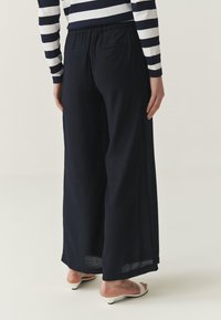 Pantalones de pierna ancha en color navy, con una tela texturizada, cinturilla elástica y dos bolsillos traseros. Combinados con sandalias deslizables blancas.