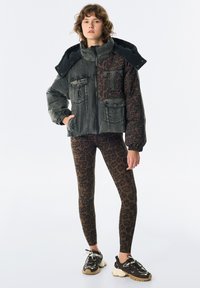 Jeune personne portant une veste en tissu mélangé avec un imprimé léopard et du denim sombre, associée à des leggings imprimés léopard brun et des baskets noires.