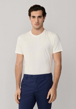 Mann mit dunklen Haaren, der ein schlichtes weißes kurzärmeliges T-Shirt und marineblaue Hosen trägt und vor einem hellgrauen Hintergrund steht.