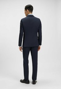 Giacca da abito blu navy con una vestibilità su misura, design a un solo petto e polsini di camicia azzurri. Indossata con pantaloni coordinati e scarpe nere.