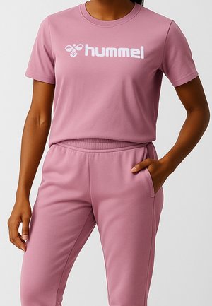 Osoba nosząca wrzosowy krótki rękaw t-shirt z logo "hummel" i dopasowane wrzosowe spodnie, ręka w kieszeni na jednolitym tle.