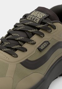 Olive green sneaker with textured material, dark gray accents, and patterned laces. Features a Vans logo on the side and tongue.
