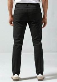 Črne hlače ozkega kroja z gladko teksturo, z dvema zadnjima žepoma in subtilnim leskom, kombinirane s svetlimi čevlji.