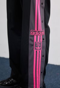 Melna un pelēka sporta bikses ar rozā svītrām, pogām uz posmām un izšūtu Adidas logotipu ar trifolijas grafiku sānos.