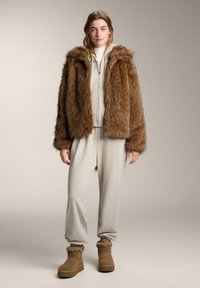 Barna műszőrme kabát kapucnival, bézs cipzáros pulóver, szürke melegítőnadrág és barna birkabőr csizma. Puha anyagok és laza szabás.