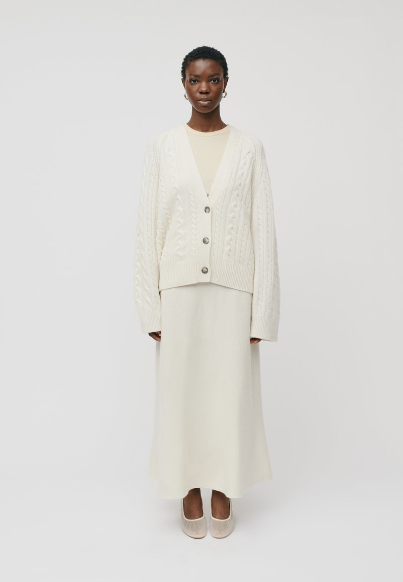 Moteris, vilkinti grietinėlės spalvos megztą kardiganą su mygtukais, atitinkančią ilgu sijonu, kreminės spalvos palaidine, aukso spalvos auskarais žiedais ir smėlio spalvos balerinomis, stovi.