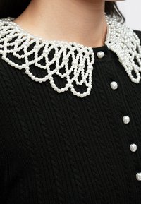 Pull en tricot noir avec des boutons en perle blanche et un col décoratif orné de perles blanches porté par une personne.