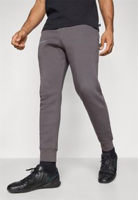 Graue Jogginghose mit schmal zulaufenden Beinen, elastischen Bündchen und einem blauen Logo-Detail. Der Stoff sieht weich aus und ist für Komfort gemacht. Getragen mit schwarzen Turnschuhen.