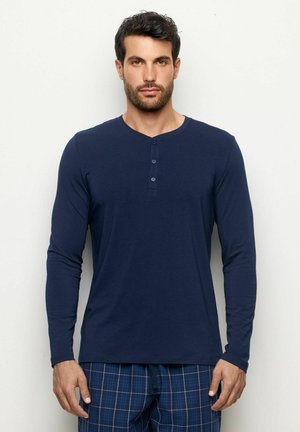 Marineblaues Langarm-Henley-Shirt mit Rundhalsausschnitt, Knopfleiste und weichem Stoff. Getragen mit karierter Schlafanzughose.