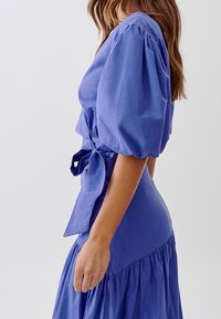Abito blu con maniche a sbuffo, vita legata, silhouette a forma di A e gonna arricciata. Tessuto morbido con una texture opaca e senza motivi visibili.