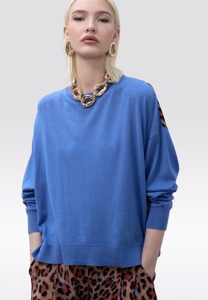 Mujer con un suéter azul holgado, collar de cadena gruesa de oro y pendientes, con pantalones de estampado de leopardo y las manos en los bolsillos.