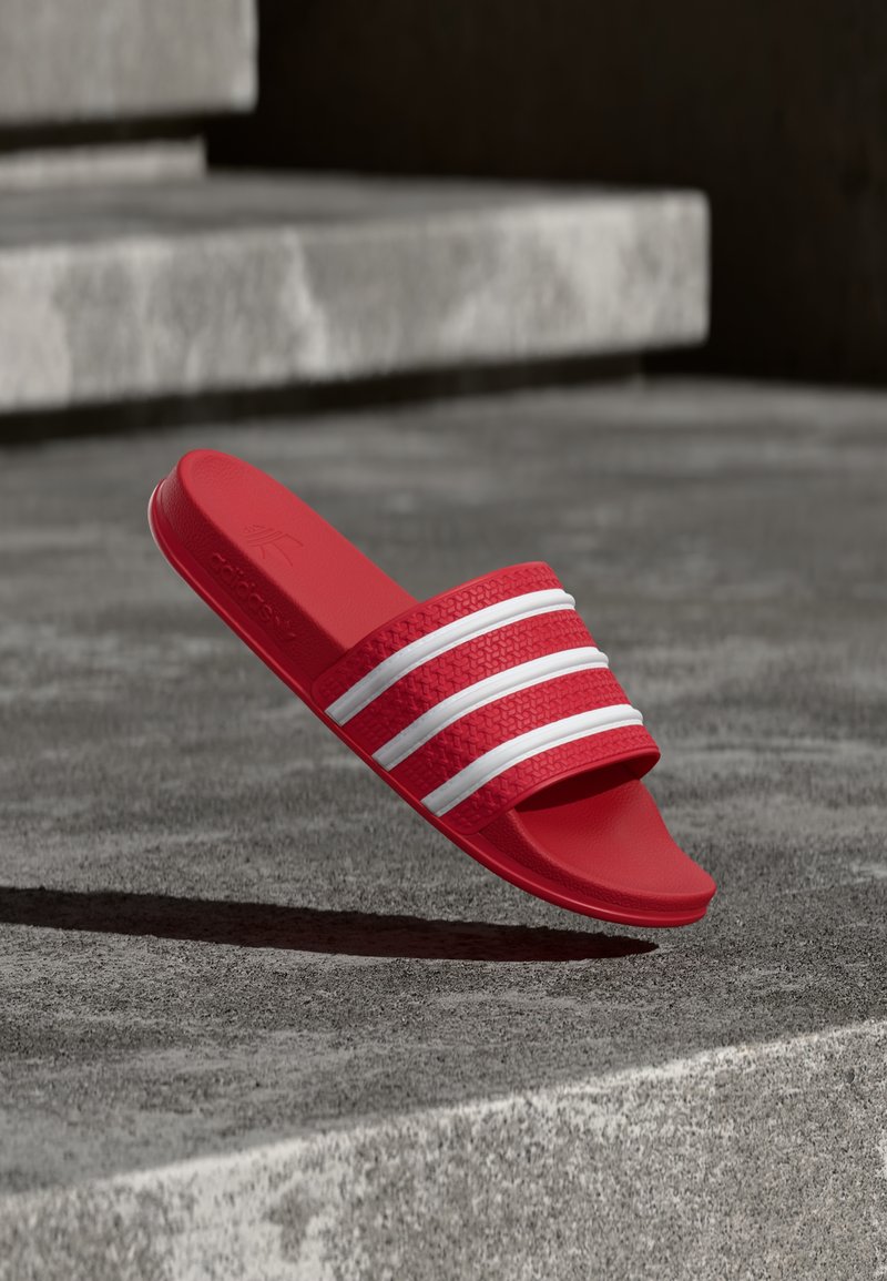 Κόκκινο σανδάλι Adidas slide με τρεις λευκές ρίγες, αιωρούμενο πάνω από γκρι σκάλες από σκυρόδεμα σε απαλές σκιές.