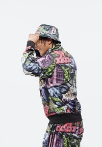 Ung person som ler, klädd i färgglad streetwear med kollage av grafiska tryck och matchande bucket-hatt, mot en enkel vit bakgrund.