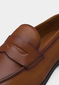 Mocassin en cuir marron avec une sangle décorative sur le dessus et un bout arrondi, présentant des coutures visibles et une semelle noire.