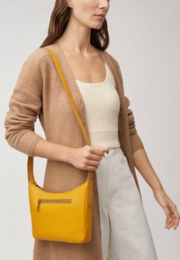 Sac à épaule en cuir jaune avec une poche zippée, texture lisse, forme douce et longue sangle, porté par une personne dans un cardigan beige.