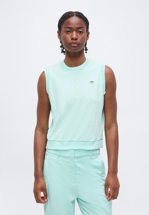 Noor inimene, kes kannab varrukateta laimirohelist mustrilist tuba ja sobivaid helerohelisi pükse, seisab ühevärvilise valge tausta ees.