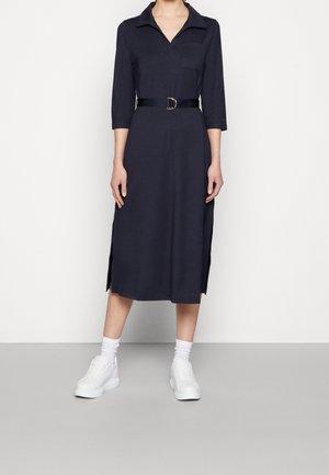 Kobieta w granatowej midi sukience koszulowej z rękawami 3/4, z rozcięciami po bokach, czarnym paskiem, białymi skarpetkami i białymi masywnymi trampkami.