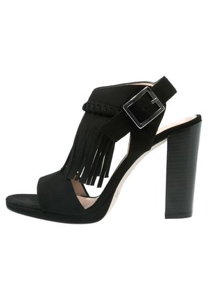 Sandale noire à talons hauts avec un bout ouvert, en suède, détail frangé et une sangle à boucle réglable. Elle présente un talon empilé.