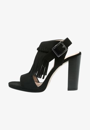 Sandale noire à talons hauts avec un bout ouvert, en suède, détail frangé et une sangle à boucle réglable. Elle présente un talon empilé.