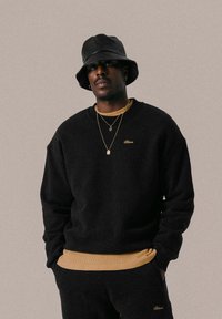 Schwarzer Rundhals-Sweatshirt mit goldener Stickerei, über einem tanfarbenen Unterhemd getragen. Kombiniert mit einem schwarzen Bucket Hat und geschichteten goldenen Halsketten.