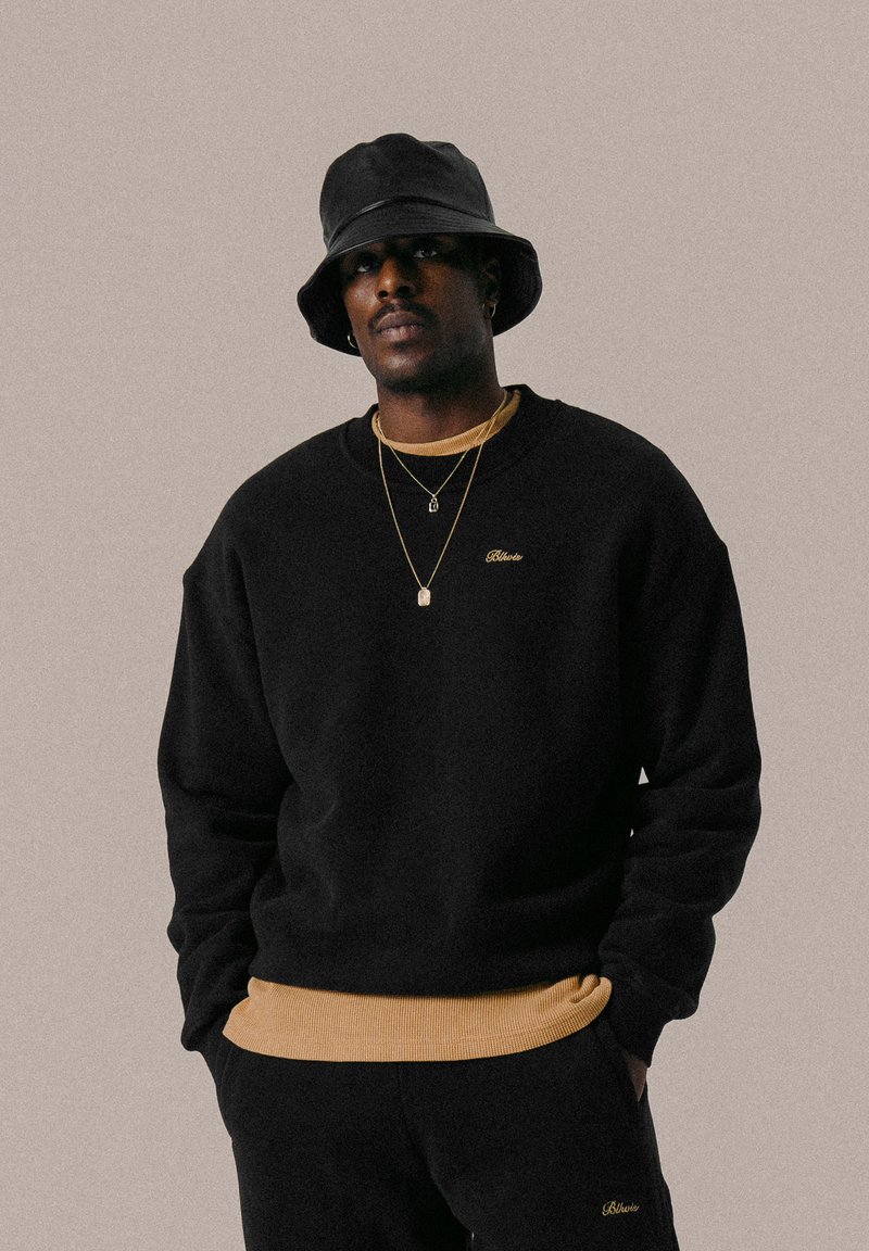 Schwarzer Rundhals-Sweatshirt mit goldener Stickerei, über einem tanfarbenen Unterhemd getragen. Kombiniert mit einem schwarzen Bucket Hat und geschichteten goldenen Halsketten.