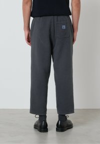 Osoba oblečená v tmavošedých voľných nohaviciach s zadnou nášivkou a čiernymi formálnymi topánkami, stojaca na svetlošedej podlahe.