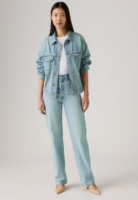 Lichtblauwe spijkerjas met voorzakken, gecombineerd met bijpassende lichtblauwe jeans. De outfit heeft een wit shirt en tan gekleurde puntige schoenen.