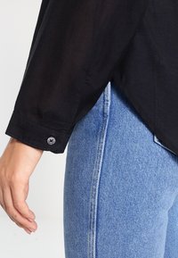 Black, long-sleeve shirt with a buttoned cuff, paired with light blue denim jeans showcasing a fitted design and a slightly tapered shape.