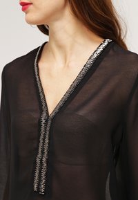 Donna che indossa una blusa nera trasparente con rifiniture argento lungo il collo a V, con un sottogonna nera senza spalline visibile sotto.