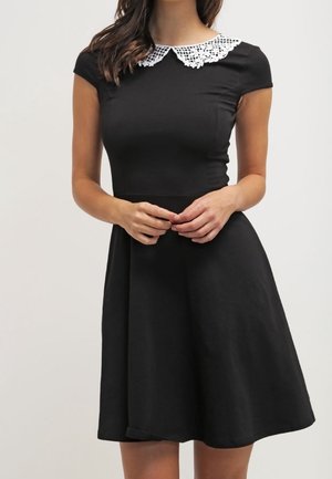Donna che indossa un vestito nero corto con corpetto aderente, maniche corte, gonna svasata e colletto Peter Pan in pizzo bianco, in piedi con le mani giunte.