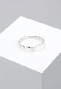 Anillo delgado de plata con una sutil hendidura en forma de V, apoyado sobre un pedestal cúbico blanco contra un fondo gris neutro.