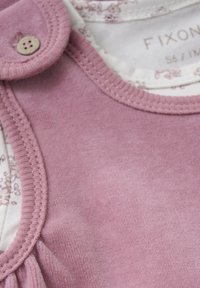 Vêtement en velours rose clair avec un col rond, présentant une fermeture à boutons et un motif floral sur le tissu intérieur. Texture lisse.