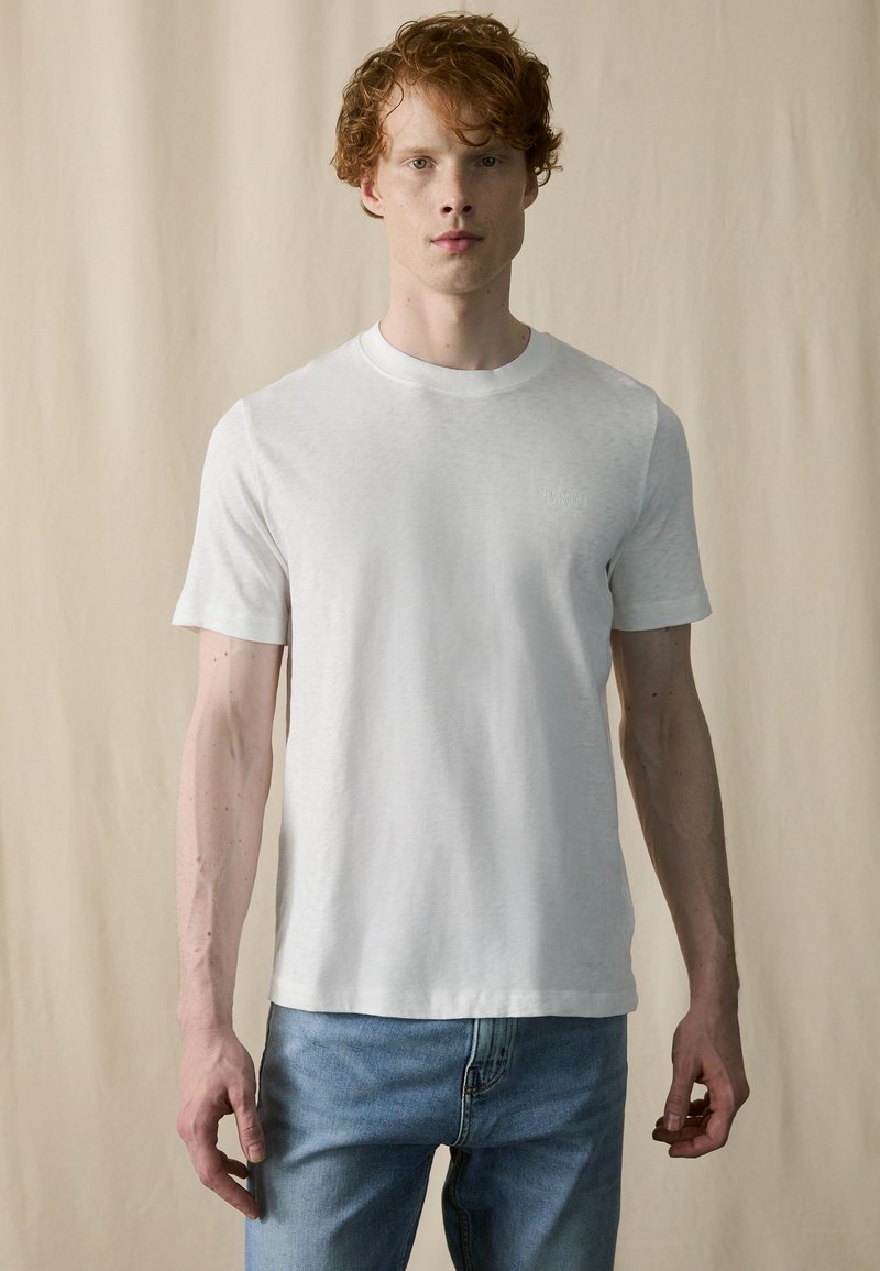 Hvit bomull T-skjorte med rund hals og korte ermer, med subtil tekstur og en liten logo på brystet. Brukt med lyseblå jeans.