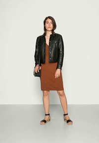 Jaqueta de couro preta sobre um vestido justo castanho, combinada com sandálias pretas de tiras. Bolsa transversal preta simples, fundo neutro.