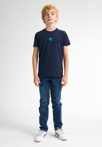 Námorně modré bavlněné tričko s kulatým výstřihem a krátkými rukávy, na přední straně s modrým grafickým logem. Oblečeno s modrými džíny a bílými teniskami.