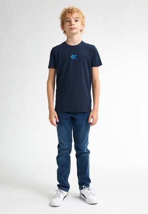 Marineblaue Baumwoll-T-Shirt mit rundem Ausschnitt und kurzen Ärmeln, ausgestattet mit einem blauen Grafiklogo auf der Brust. Kombiniert mit blauen Jeans und weißen Sneakers.