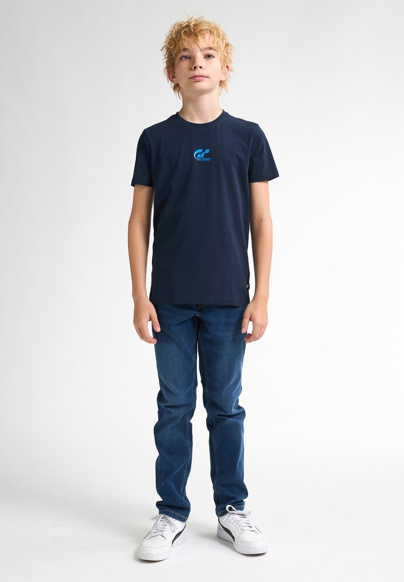 Námorně modré bavlněné tričko s kulatým výstřihem a krátkými rukávy, na přední straně s modrým grafickým logem. Oblečeno s modrými džíny a bílými teniskami.