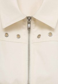 Cremefarbener Blazer mit Reißverschluss, silberner Hardware und Ösen-Details. Verfügt über einen spitz zulaufenden Kragen und eine glatte Stofftextur.