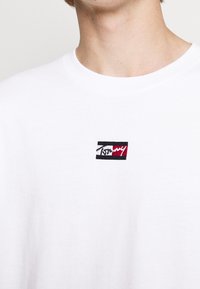 Nahaufnahme einer Person, die ein weißes Rundhals-T-Shirt mit einem kleinen rechteckigen roten, weißen und schwarzen Logo auf der Brust trägt.
