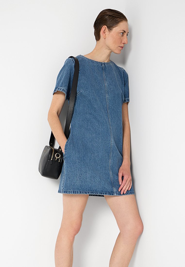 Robe chemise en denim bleu avec des manches courtes, un col rond et des coutures visibles. Associée à un petit sac à bandoulière noir.