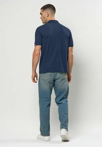 Férfi sötétkék rövid ujjú pólóban, világoskék farmerben és fehér tornacipőben, háttal áll egy egyszerű világos háttér előtt.