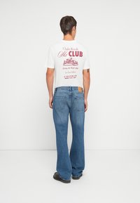 Hvit t-skjorte med rødt grafisk trykk på baksiden, med skiklubb-tema. Mørkeblå utsvingte jeans med fem lommer og et brunt lærmerke.