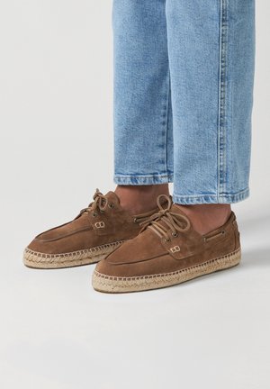 Hnědé semišové příležitostné boty s pletenými jutovými podrážkami, s šněrovacím designem a kovovými očky. Vytvořené v kombinaci se světle modrými džíny s ohrnutými nohavicemi.