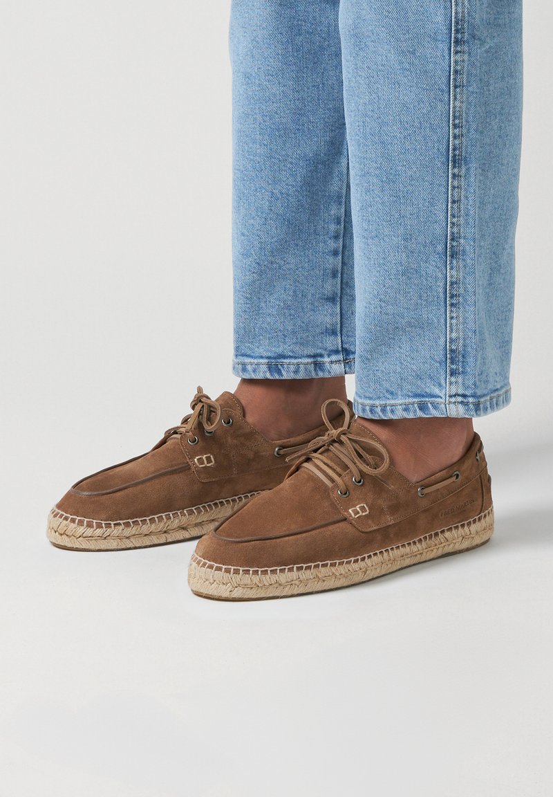 Chaussures décontractées en daim marron avec des semelles en jute tressé, à lacets et avec des œillets en métal. Portées avec un jean léger bleu avec revers.