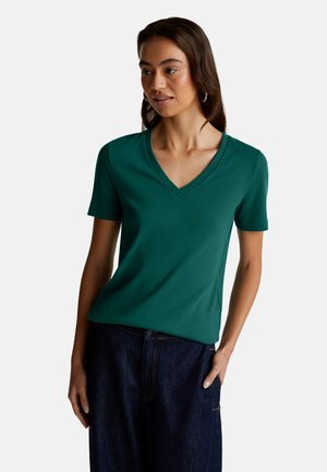 Woman wearing a green V-neck short sleeve shirt and dark blue jeans, standing with one hand in pocket against a plain background.