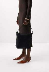 Czarna skórzana torba na ramię o strukturalnym kształcie, z paskiem na ramię i srebrnym logo. Osoba ubrana w brązową sukienkę z prążkowanego materiału i brązowe czółenka ze spiczastym noskiem.