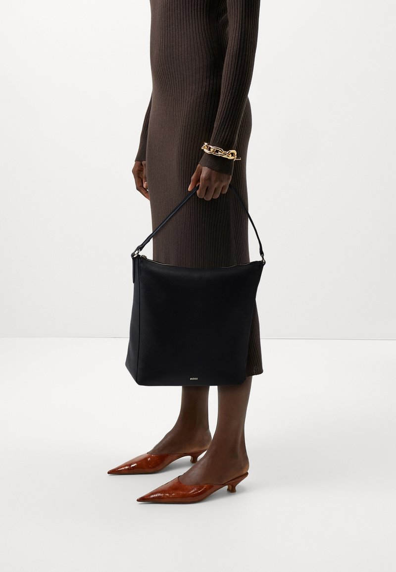 Czarna skórzana torba na ramię o strukturalnym kształcie, z paskiem na ramię i srebrnym logo. Osoba ubrana w brązową sukienkę z prążkowanego materiału i brązowe czółenka ze spiczastym noskiem.