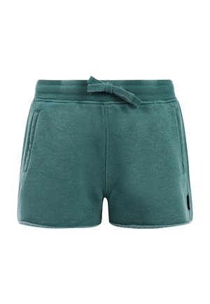 Lachend meisje met lang haar dat een zwart shirt met korte mouwen draagt, turquoise shorts met een trekkoord en zwarte slippers, staand met één hand in de zak.