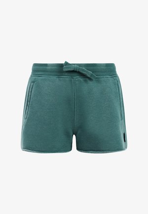 Lachend meisje met lang haar dat een zwart shirt met korte mouwen draagt, turquoise shorts met een trekkoord en zwarte slippers, staand met één hand in de zak.